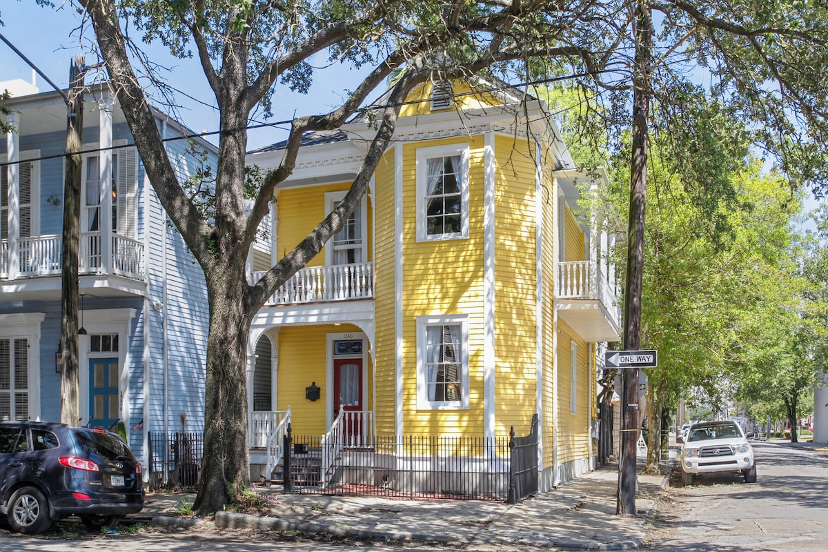 A charming yellow Victorian home is framed by lush trees, creating an inviting street view. The house features white shutters and a red front door, complemented by classic architectural details such as a gabled roof and decorative trim. Two balconies can be seen above the entrance.