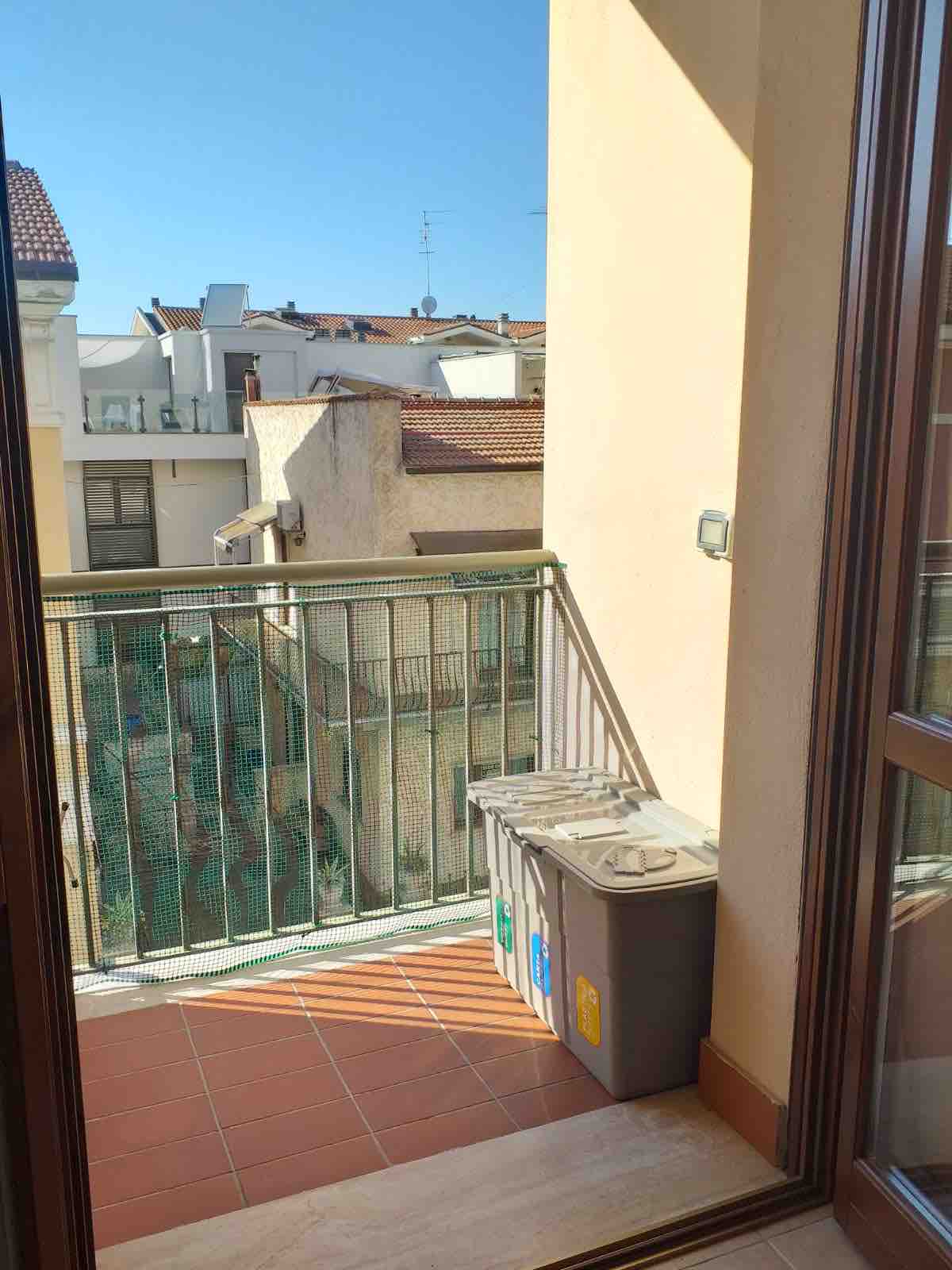 A small balcony is visible featuring a railing and tiled flooring. Waste bins are positioned against the wall, with sunlight casting shadows across the space. Surrounding buildings are seen in the background, contributing to the urban setting.