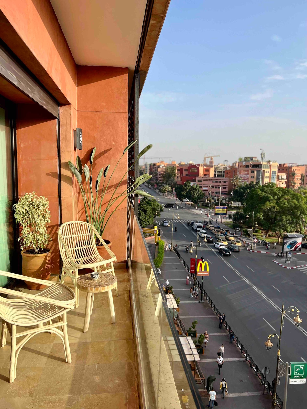 A balcony is featured with two woven chairs positioned next to a potted plant. The view overlooks a busy street with passing vehicles and buildings in the distance. Bright natural light illuminates the outdoor space, highlighting the warm tones of the surroundings.