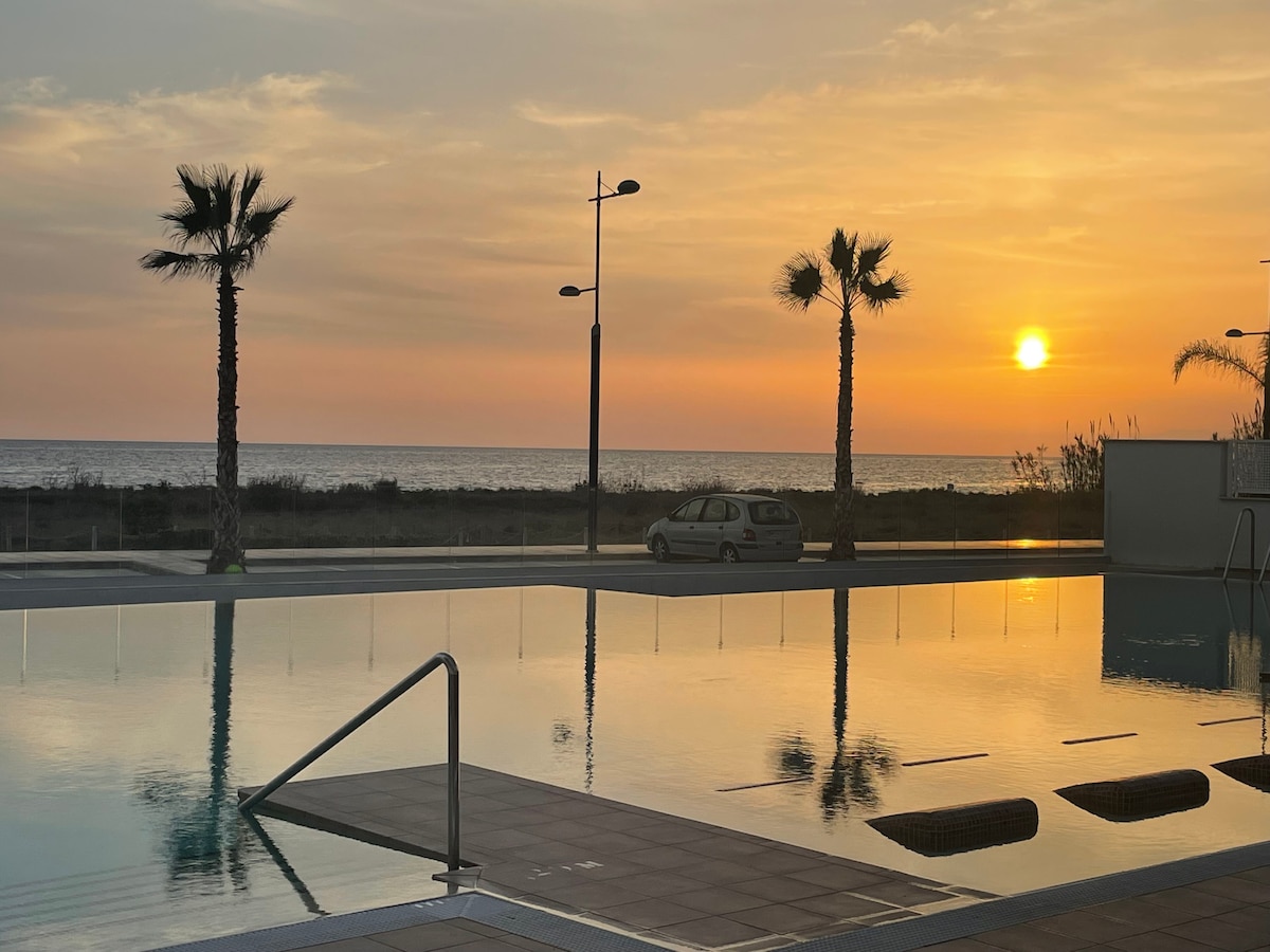 A serene sunset view is captured over the tranquil pool, with silhouettes of palm trees framing the scene. The reflection of warm hues illuminates the water surface, while the distant horizon showcases the transitioning sky and the calm sea beyond.