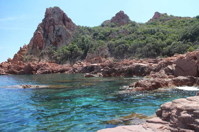 A serene coastal scene is captured, showcasing clear turquoise waters gently lapping against rocky shores. The rugged cliffs, adorned with lush greenery, rise prominently in the background, creating a natural barrier along the coastline.