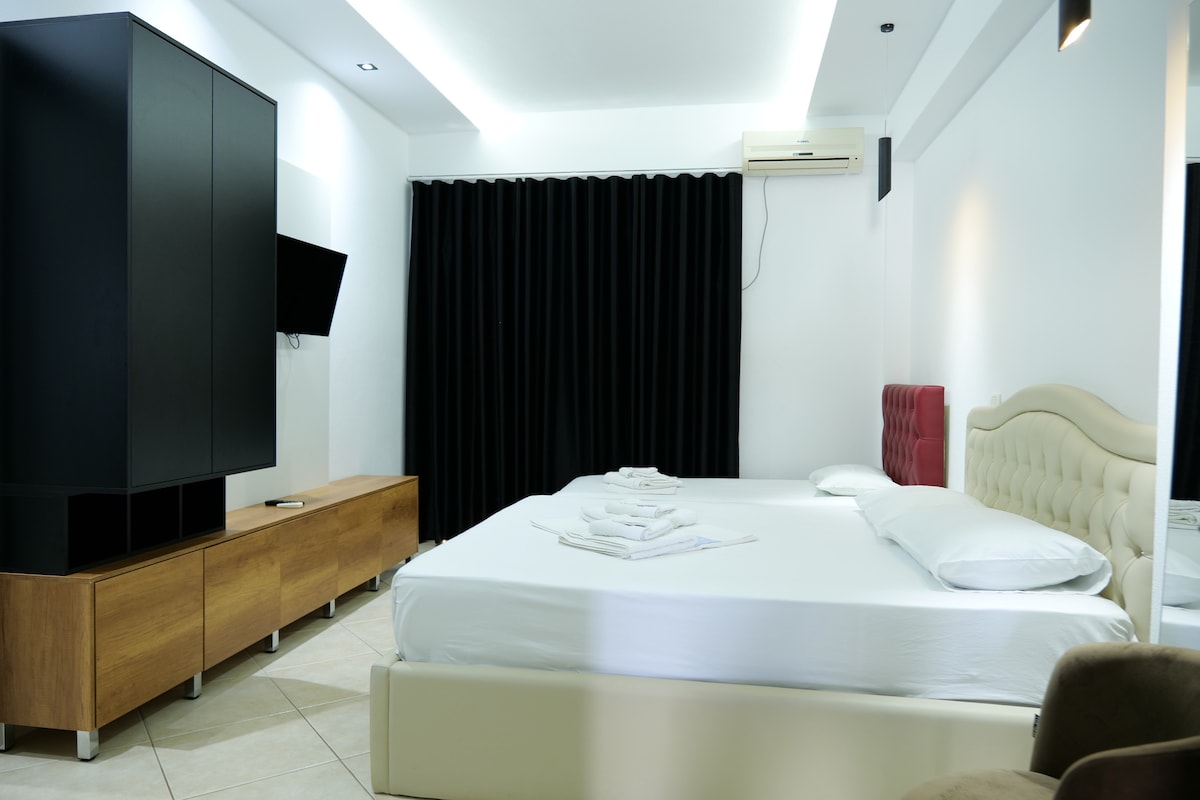 A spacious bedroom is featured with a light-colored bed and soft bedding. Dark curtains cover the window behind the bed, while a television is mounted on the wall. A sleek black wardrobe and wooden storage unit add to the modern aesthetic of the room.