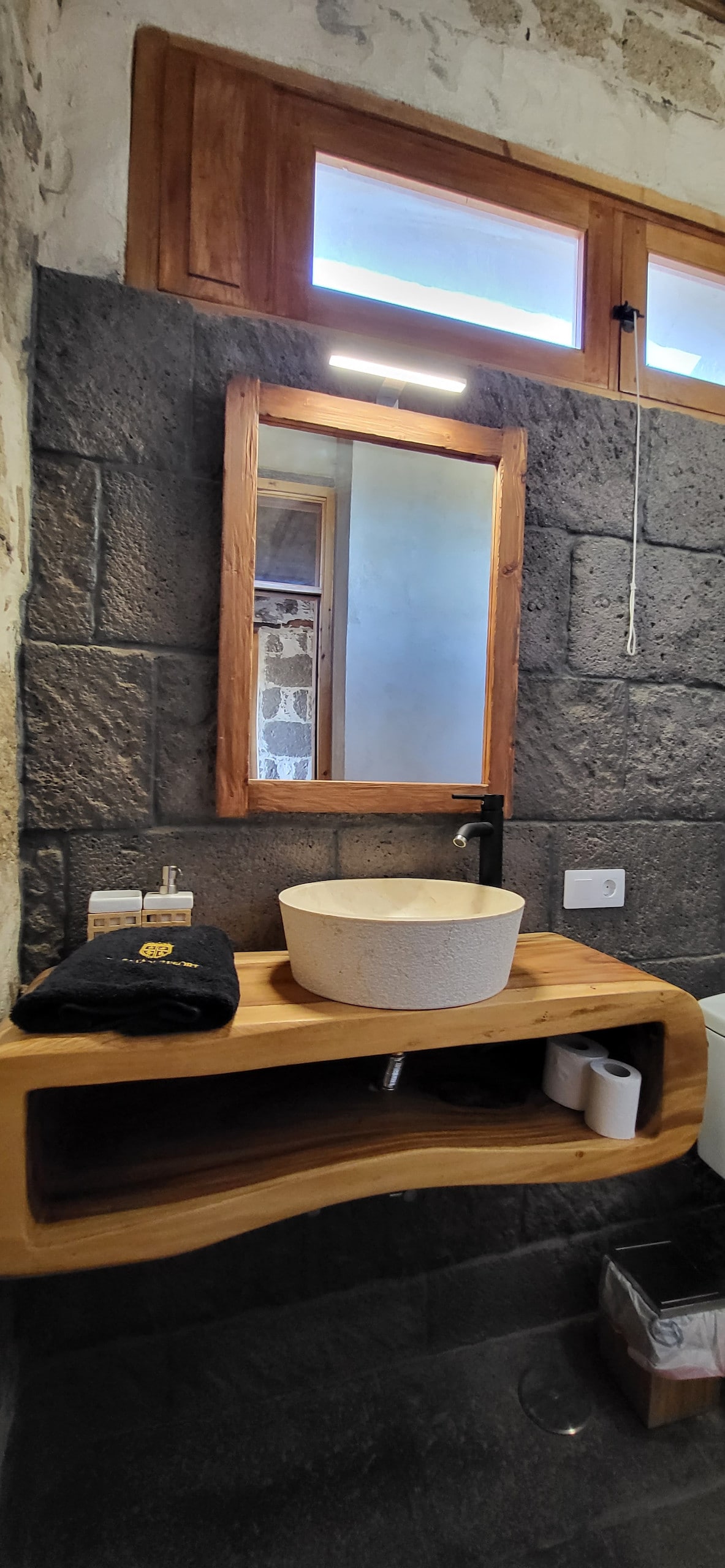 A modern bathroom features a stone wall and wooden accents. A round sink sits on a wooden countertop, with a large mirror above it. Natural light filters through the small windows, highlighting the clean lines and minimalist design of the space.