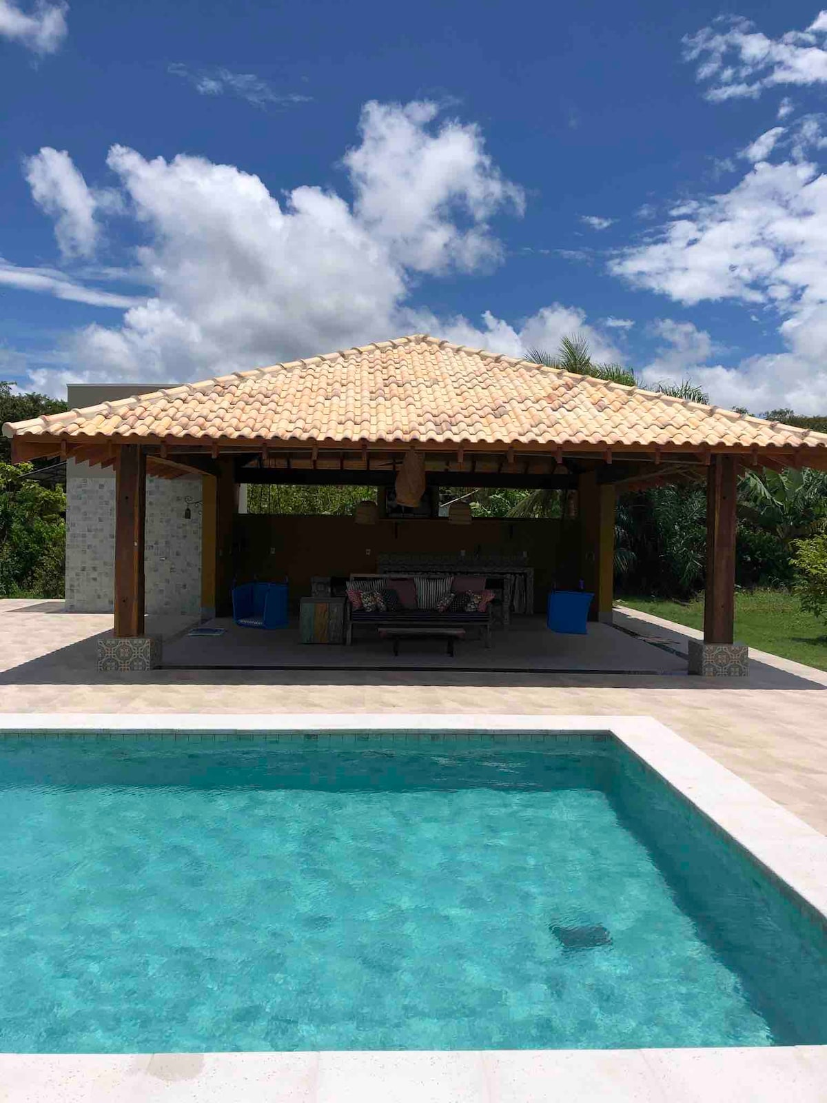 An outdoor area features a thatched-roof pavilion with a spacious seating arrangement and an outdoor kitchen. A clear, inviting pool lies in the foreground, reflecting the blue sky and surrounding greenery.
