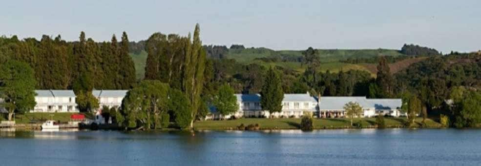 A tranquil lakeside view is presented, featuring a series of white buildings with blue roofs. Lush greenery and tall trees line the shoreline, while gentle hills rise in the background, all reflected in the calm lake water.
