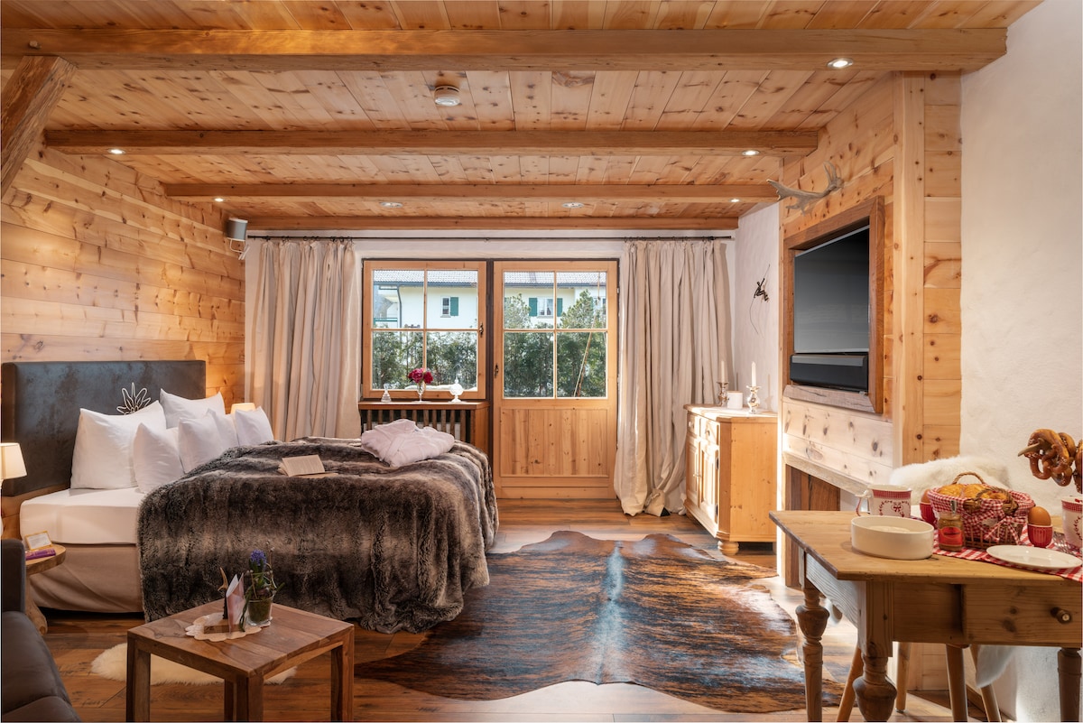 A cozy room featuring wooden walls and beams, creating a warm ambiance. A spacious bed is draped with a soft, textured blanket. Natural light fills the space through a large window, complemented by elegant curtains. A wooden table and dresser add to the rustic charm.