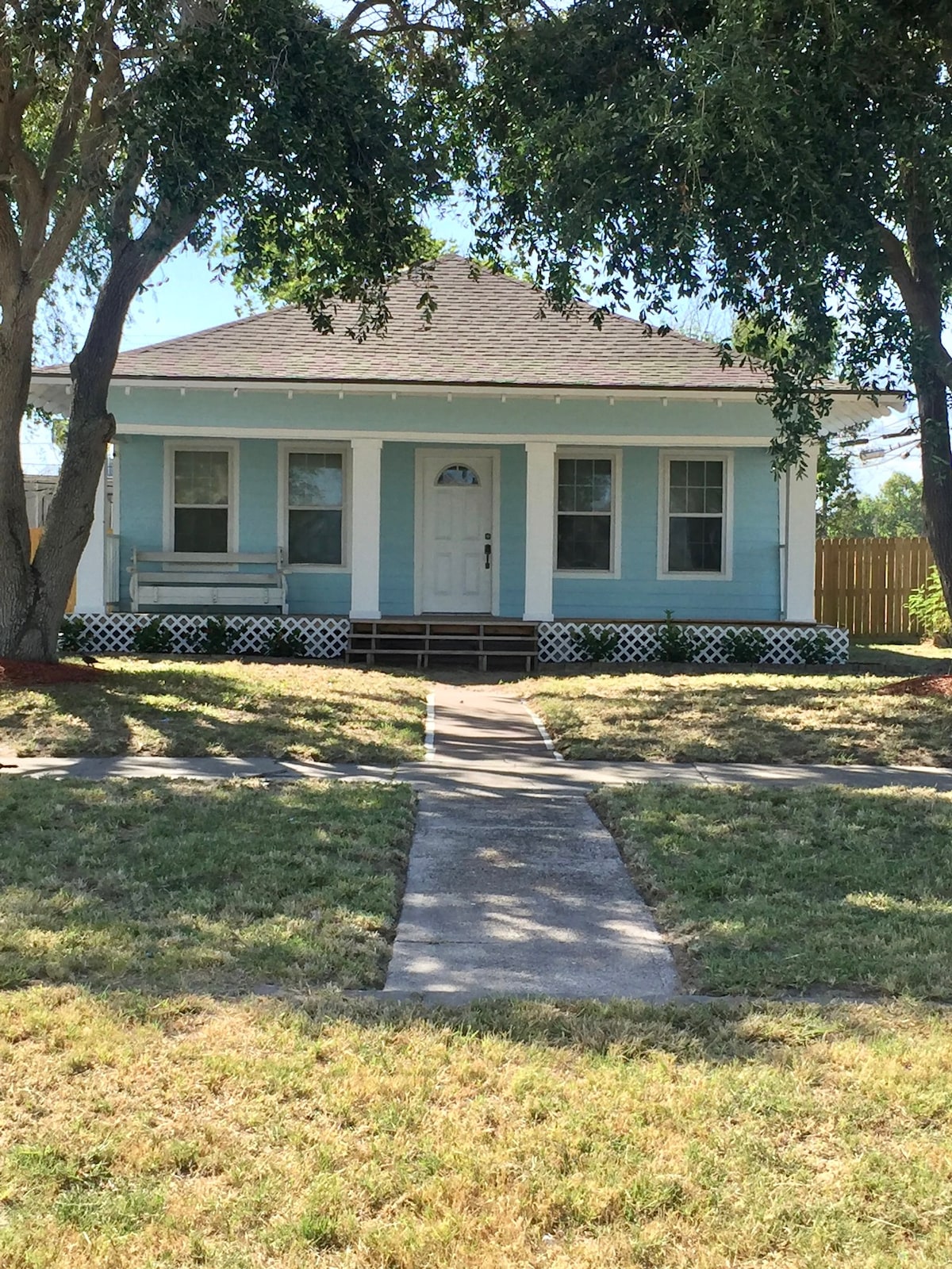 A charming single-story house features light blue exterior siding and a welcoming front porch. A path leads to the central white door, flanked by large windows. Surrounding greenery and a fenced yard create an inviting atmosphere, enhancing the home’s approachability.