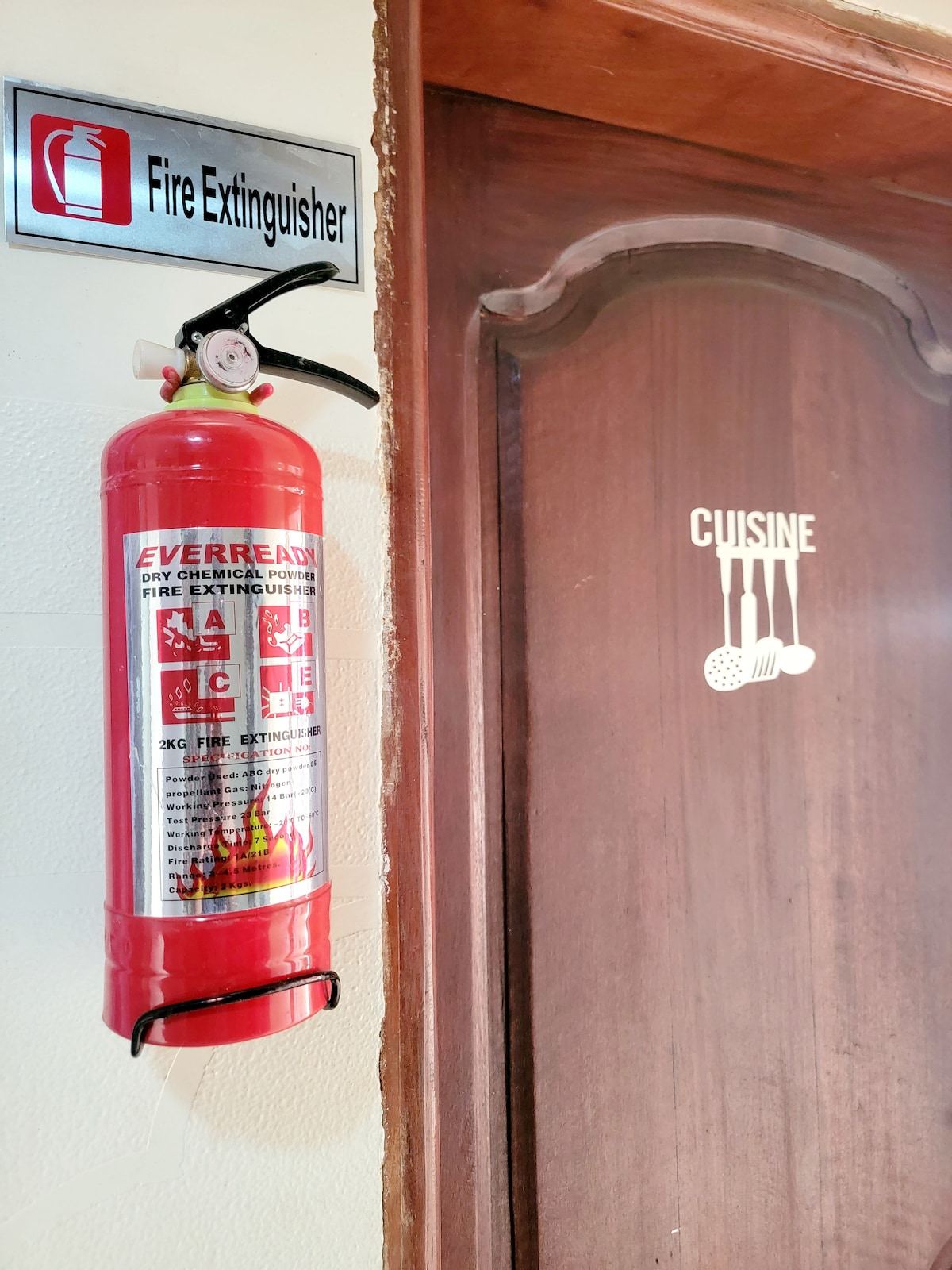 A fire extinguisher is mounted on the wall beside a wooden door. The door is labeled with the word 'Cuisine,' indicated by simple graphic utensils. The sign above the extinguisher displays a symbol for fire safety, contributing to the overall safety features of the space.