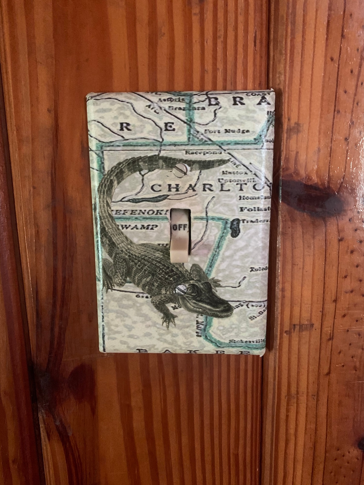 A light switch cover, decorated with a vintage map design featuring a reptile illustration, is mounted on a heart-pine wall. The texture of the wood is visible, highlighting the rustic charm of the cabin.