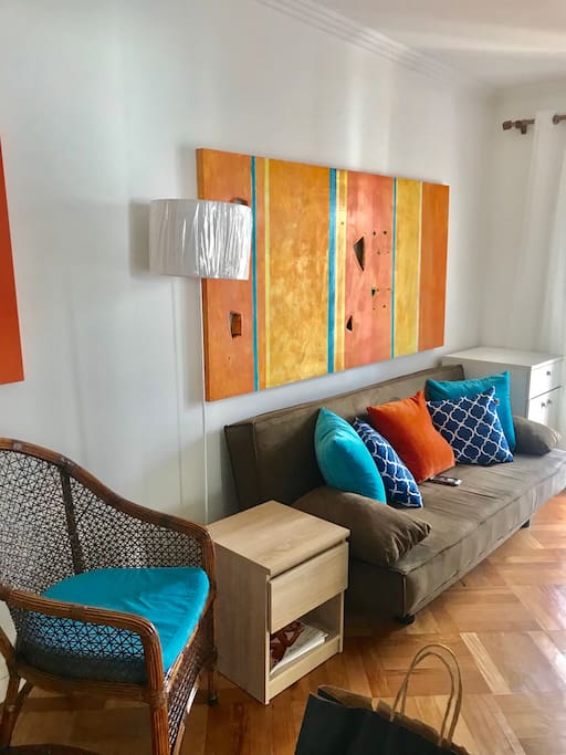 Vista da sala d jantar e tv luminaria tela em tons d laranja , mesinha apoio, almofadas laranja e azuis , janela cortina em voile e algodao e poltrona de palhinha c assento em azul claro