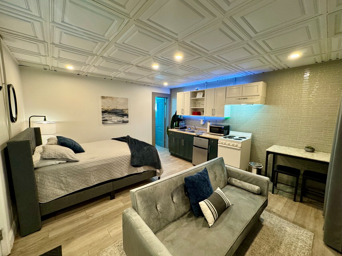 A studio space is presented featuring a spacious bed with neutral bedding and a cozy gray sofa. The kitchen area includes modern appliances with under-cabinet lighting, while a dining table for two provides a functional space. The room is illuminated by overhead lights and natural light.