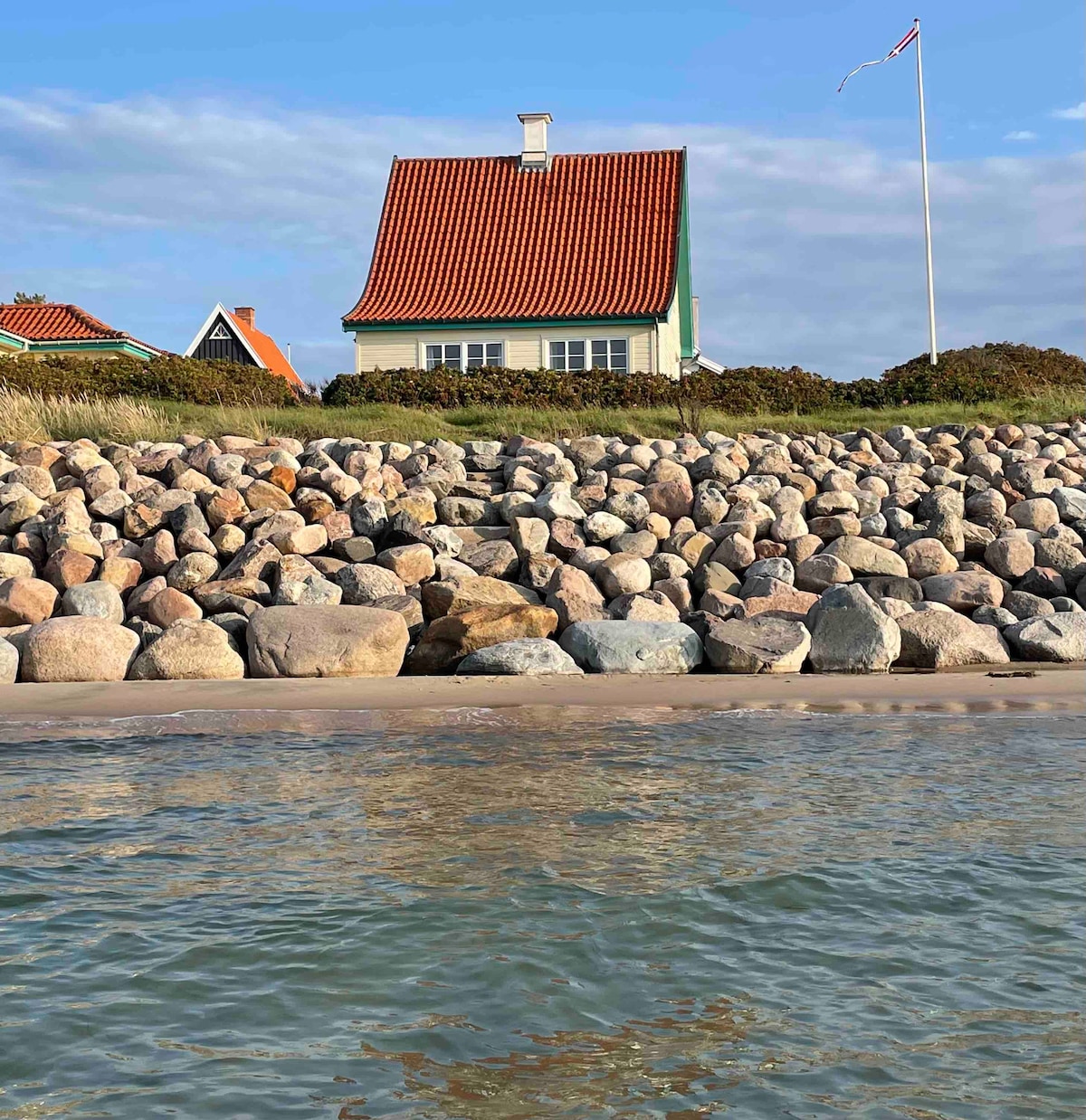 The image displays a charming house with a red-tiled roof, set atop a stone embankment. The sea is visible in the foreground, with gentle waves lapping at the sandy shore. A clear sky with scattered clouds creates a serene backdrop.