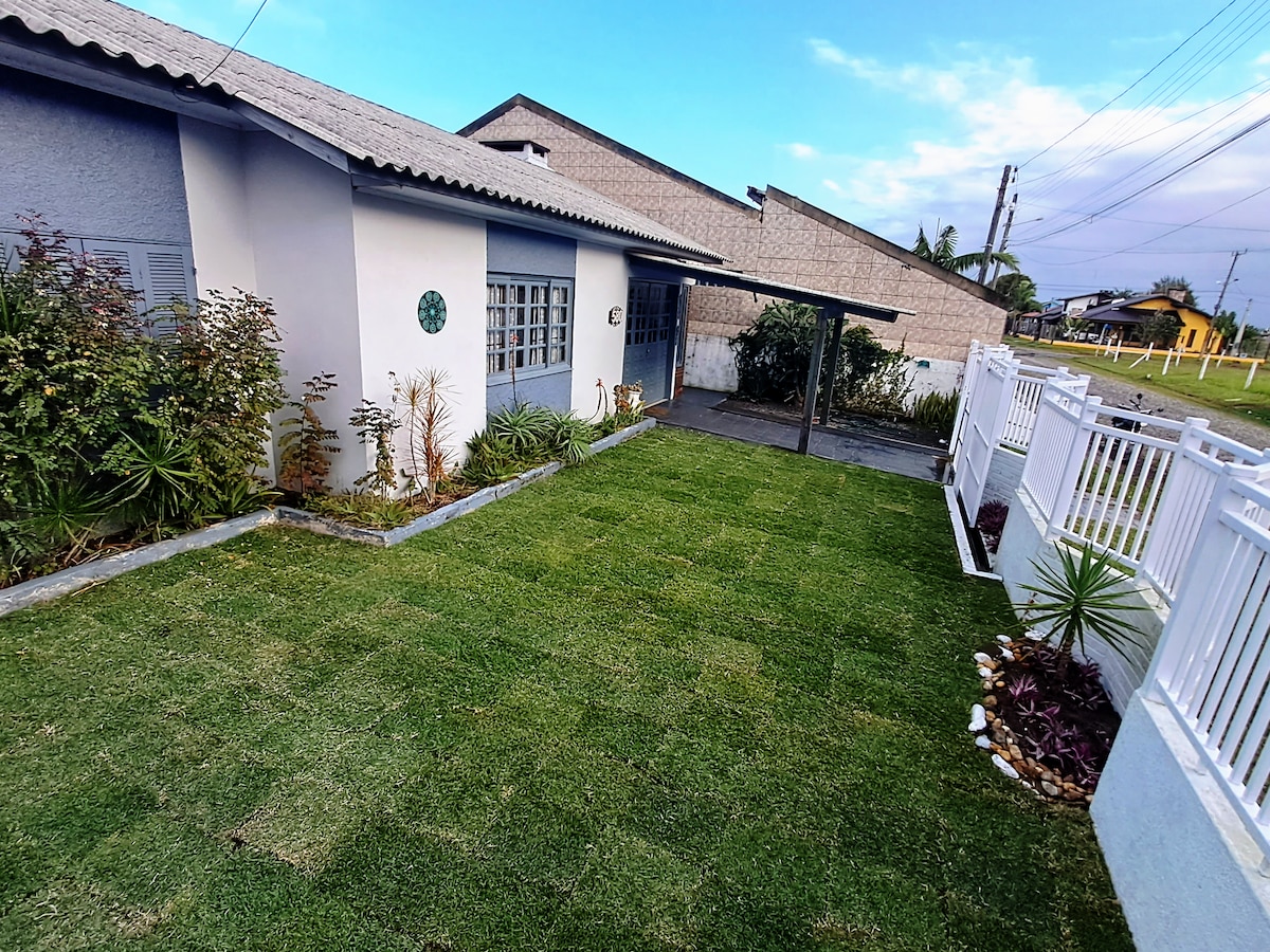 A well-maintained lawn features neatly trimmed grass surrounded by a variety of plants. The house's exterior is complemented by light-colored walls and large windows. A covered area provides shade, enhancing the outdoor space for relaxation and enjoyment.