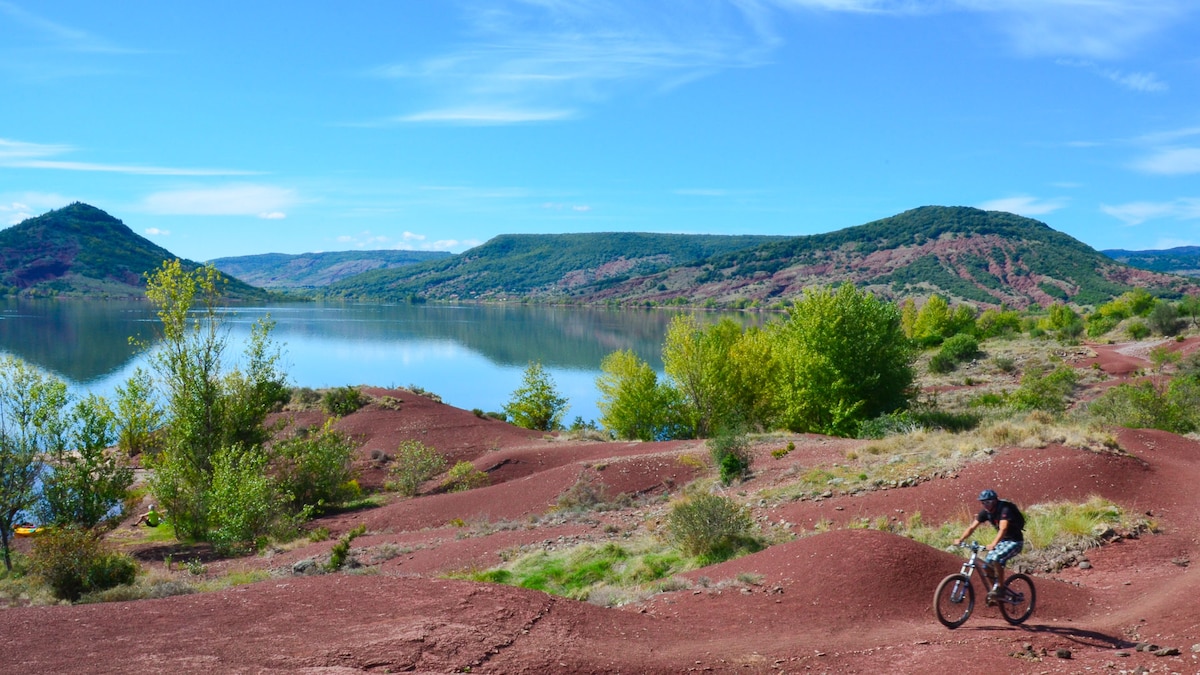 A vibrant landscape features a cyclist navigating a rugged, reddish terrain near a calm lake. Lush green trees are dispersed throughout the area, while rolling hills rise gently in the background under a clear blue sky.