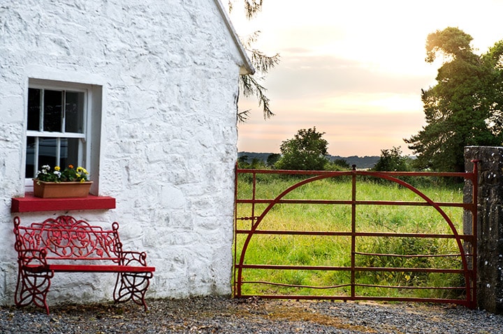 A charming exterior of the cottage features white stone walls and a vibrant red gate. A decorative bench is positioned beside the gate, complemented by a window adorned with a flower box. Lush green fields extend in the background, illuminated by the setting sun.