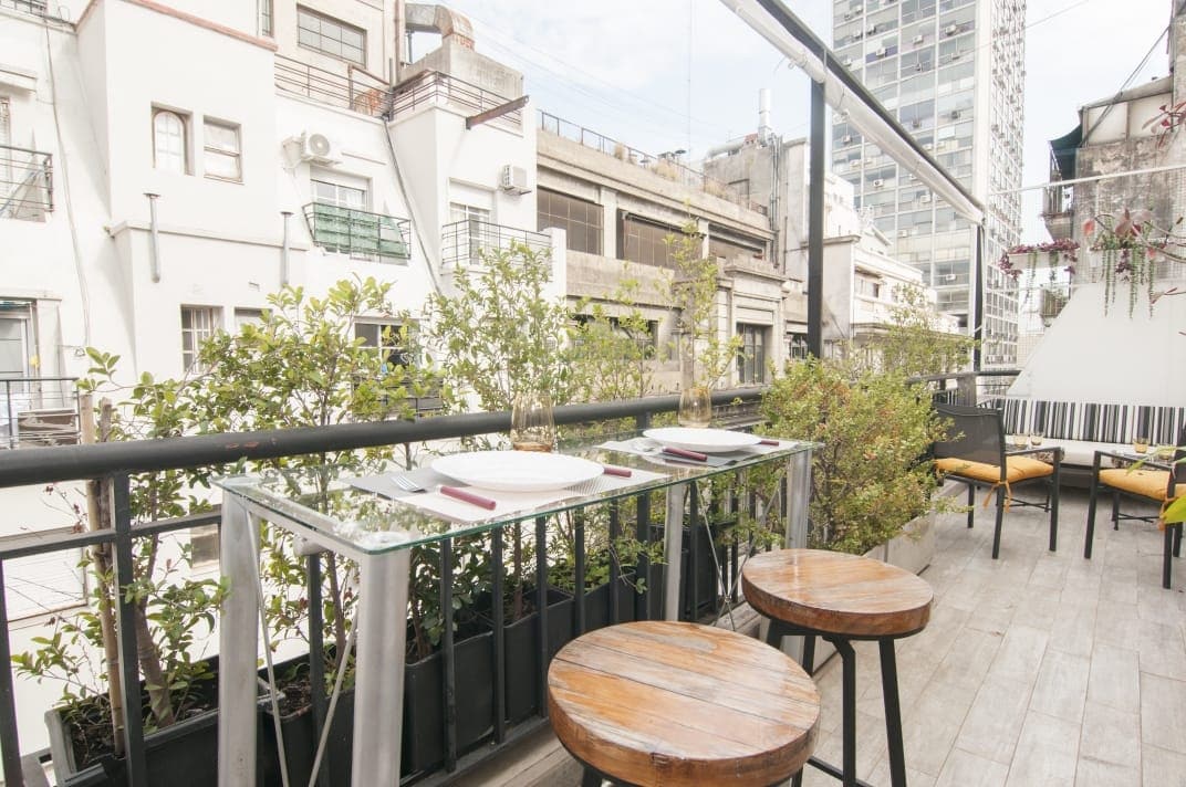 A balcony space is visible, featuring a glass-top table set for dining and two wooden stools. Green plants line the edge of the balcony, with a backdrop of surrounding buildings that provide a central urban atmosphere.