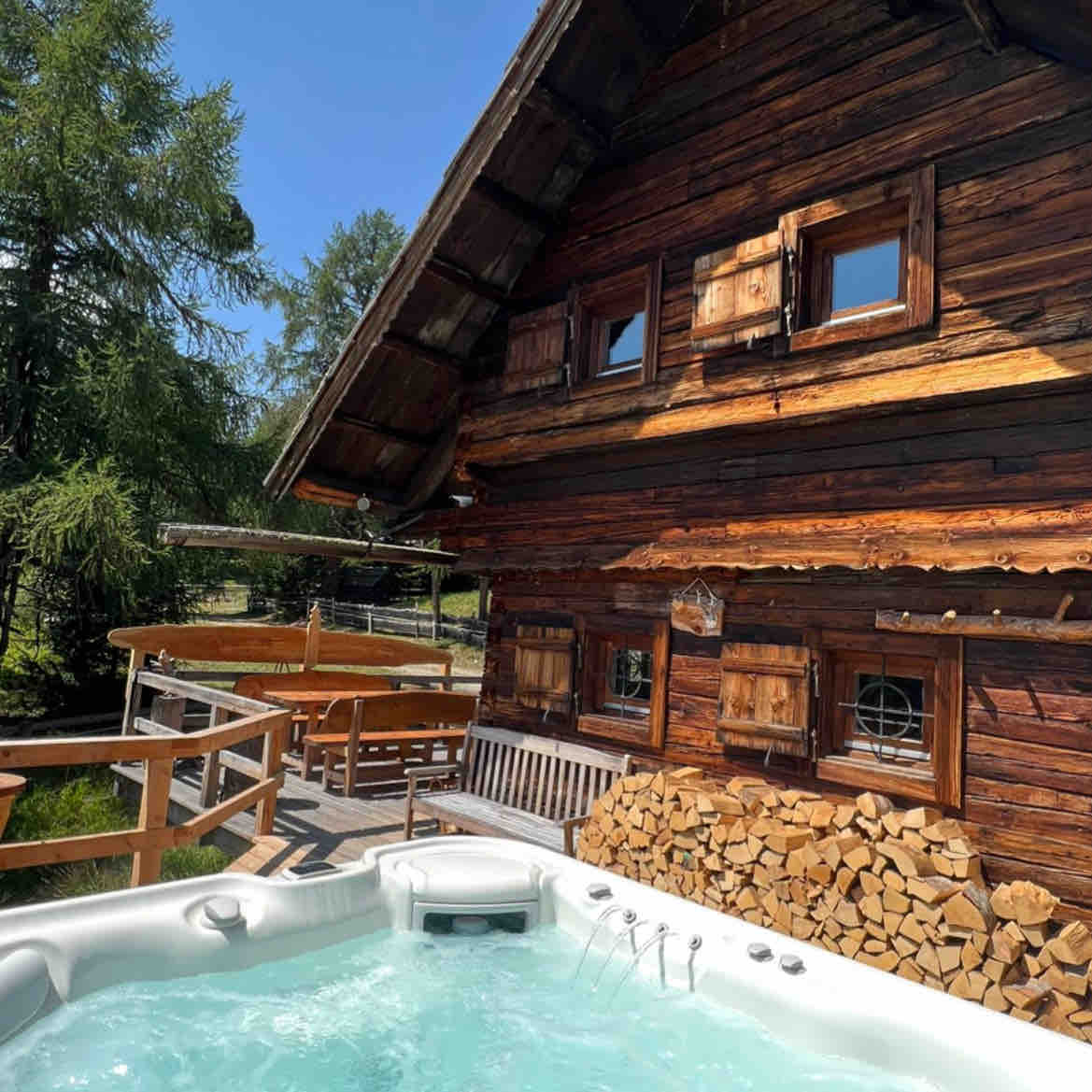 An outdoor area features a private whirlpool, surrounded by a wooden deck and rustic wooden walls of the cabin. A sitting area is visible, complemented by neatly stacked firewood nearby. Sunshine illuminates the space, enhancing the natural wooden textures.