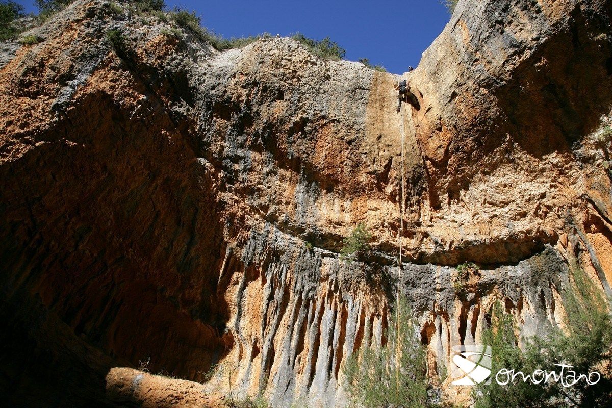The image captures a dramatic canyon with steep rock walls displaying rich earthy tones. Sunlight illuminates the textured surfaces, while climbers are visible ascending the vertical cliff. Sparse greenery is present along the ledges, contrasting with the rugged landscape creating a striking natural backdrop.
