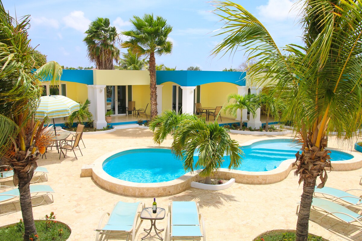 The outdoor area features a vibrant pool surrounded by palm trees and lounge chairs. A spacious terrace accommodates several seating arrangements, with a backdrop of colorful architecture under a clear blue sky.