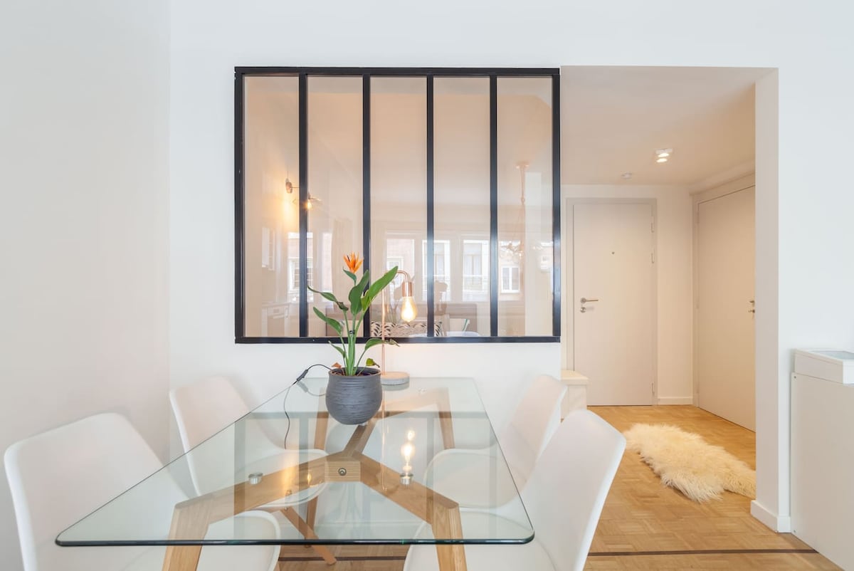 A modern dining area features a glass table with a wooden base surrounded by six white chairs. A potted plant adds a touch of greenery. The space is divided functionally from the living area, with large windows allowing natural light to filter through.