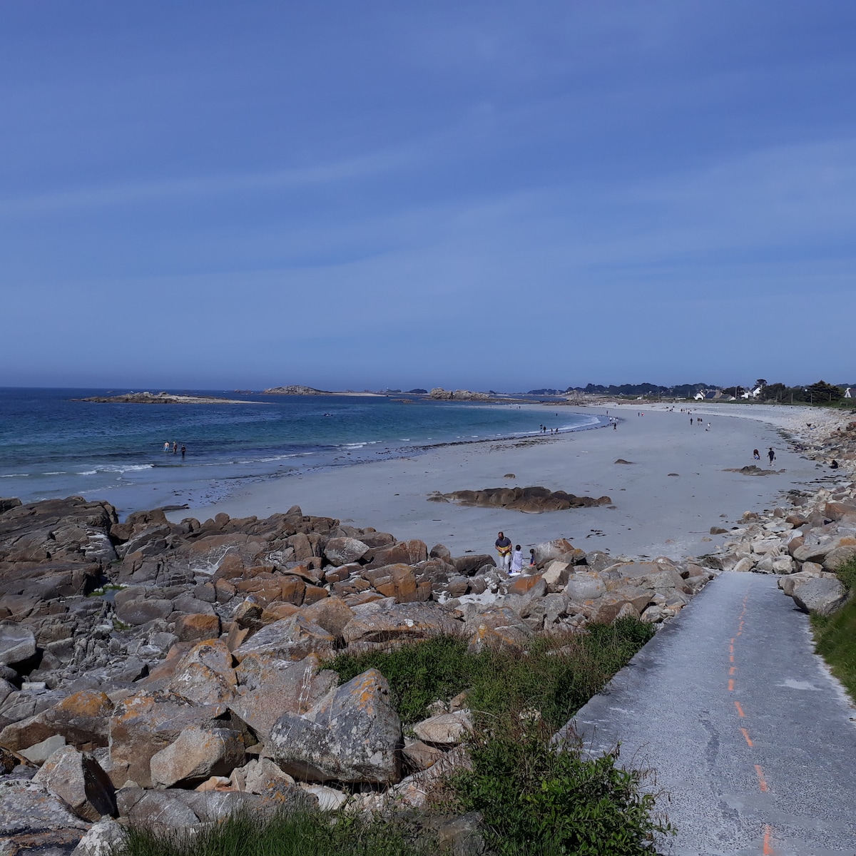 A sandy beach stretches along the coastline, framed by rocky formations and gentle waves. Visitors can be seen walking along the shore, with the ocean meeting the sky in a calm blue expanse. The scene captures a serene atmosphere, ideal for beach activities.