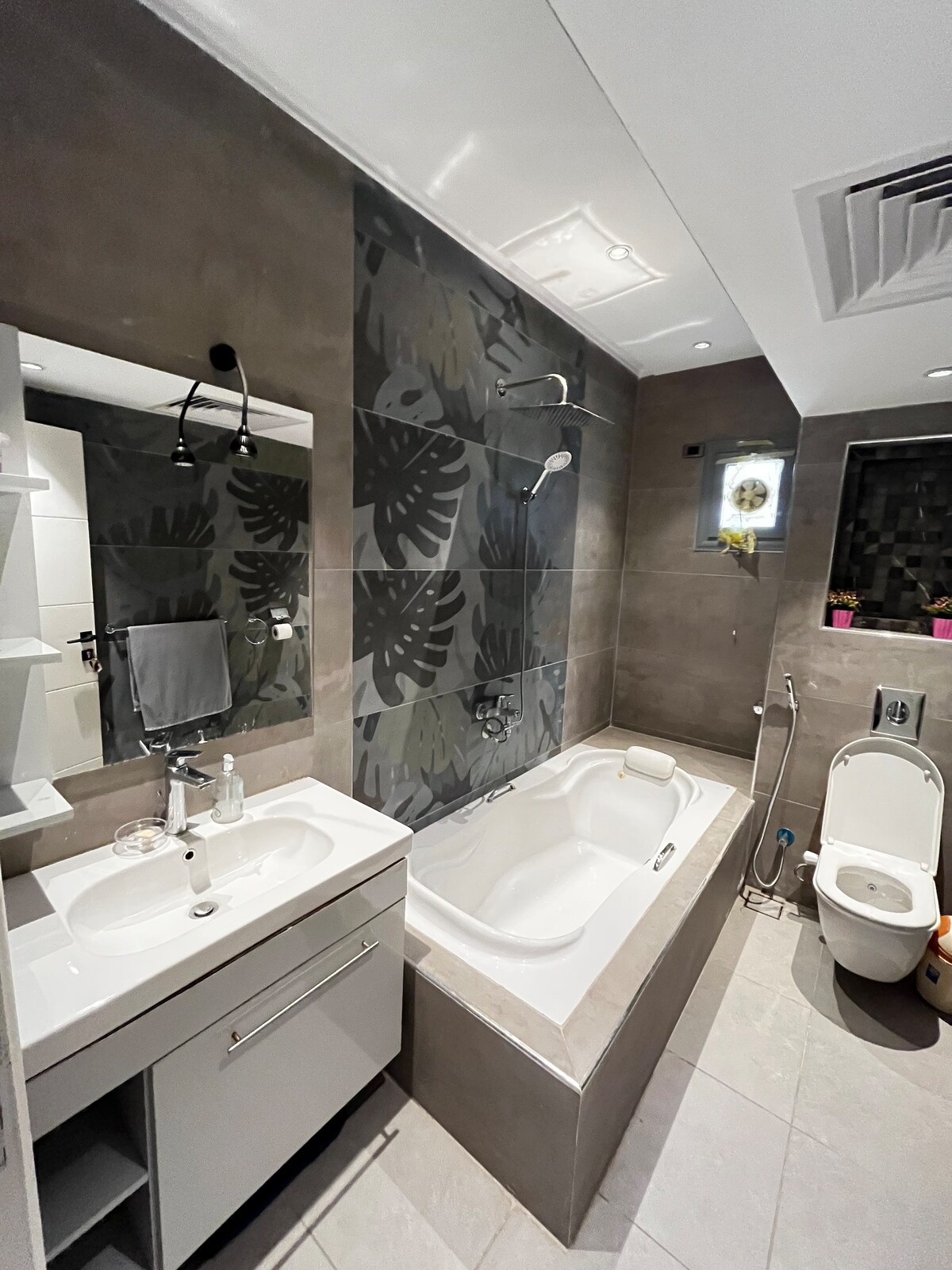A modern bathroom is featured, showcasing a sleek bathtub adjacent to a clean, white sink with storage. Large tiles with a botanical pattern create a stylish backdrop. A clear glass shower fixture and a toilet are present, complementing the neutral color scheme.
