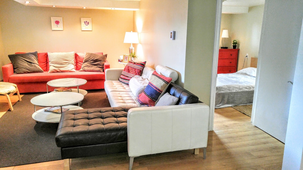 A spacious living area features a large sectional sofa in black and white, complemented by a red and a light-colored sofa. A round coffee table is positioned centrally on a dark carpet, while a soft glow emanates from the lamps nearby. An adjoining bedroom is visible, furnished with a red bed.