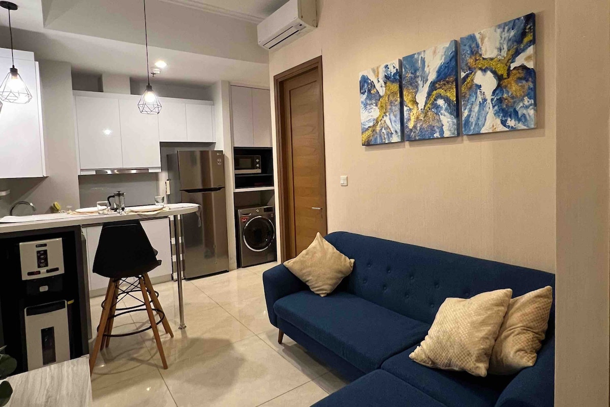 A modern living space is displayed with a blue sofa adorned with cream cushions. A dining area features a small round table with sleek stools, while wall art in vibrant blue and gold adds a pop of color. White cabinetry and sleek appliances are visible in the kitchen area.