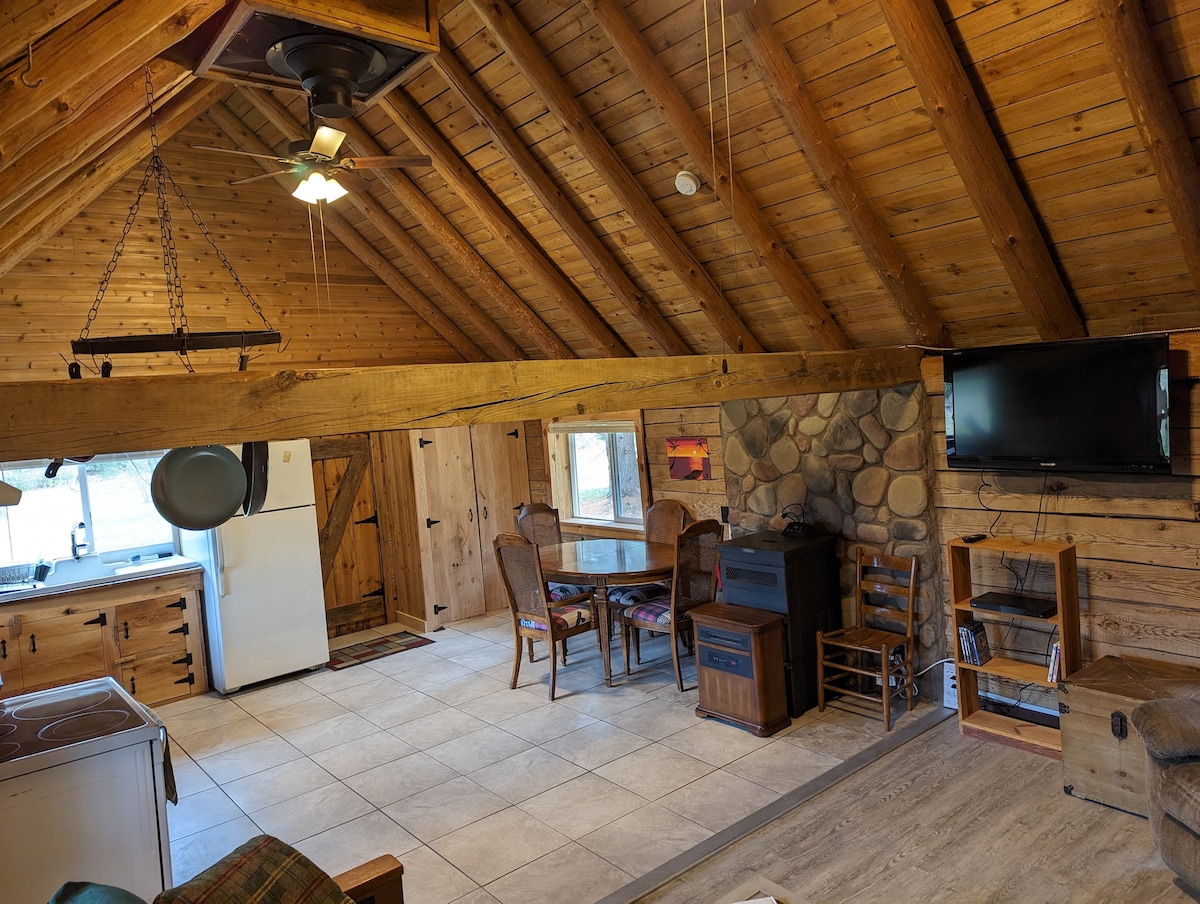An open living area is highlighted by a high, vaulted ceiling with exposed wooden beams. A circular dining table with chairs is positioned near a large window, allowing natural light. A stone wall surrounds a television mounted above a small fireplace, adding a cozy element.