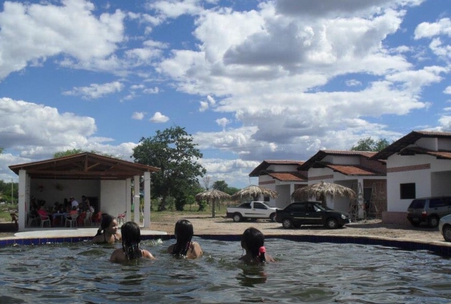 A view from the pool features three individuals enjoying the water, while a relaxation area with a roof is visible in the background. The well-maintained chalets are positioned nearby, surrounded by lush greenery and an open sky dotted with clouds.