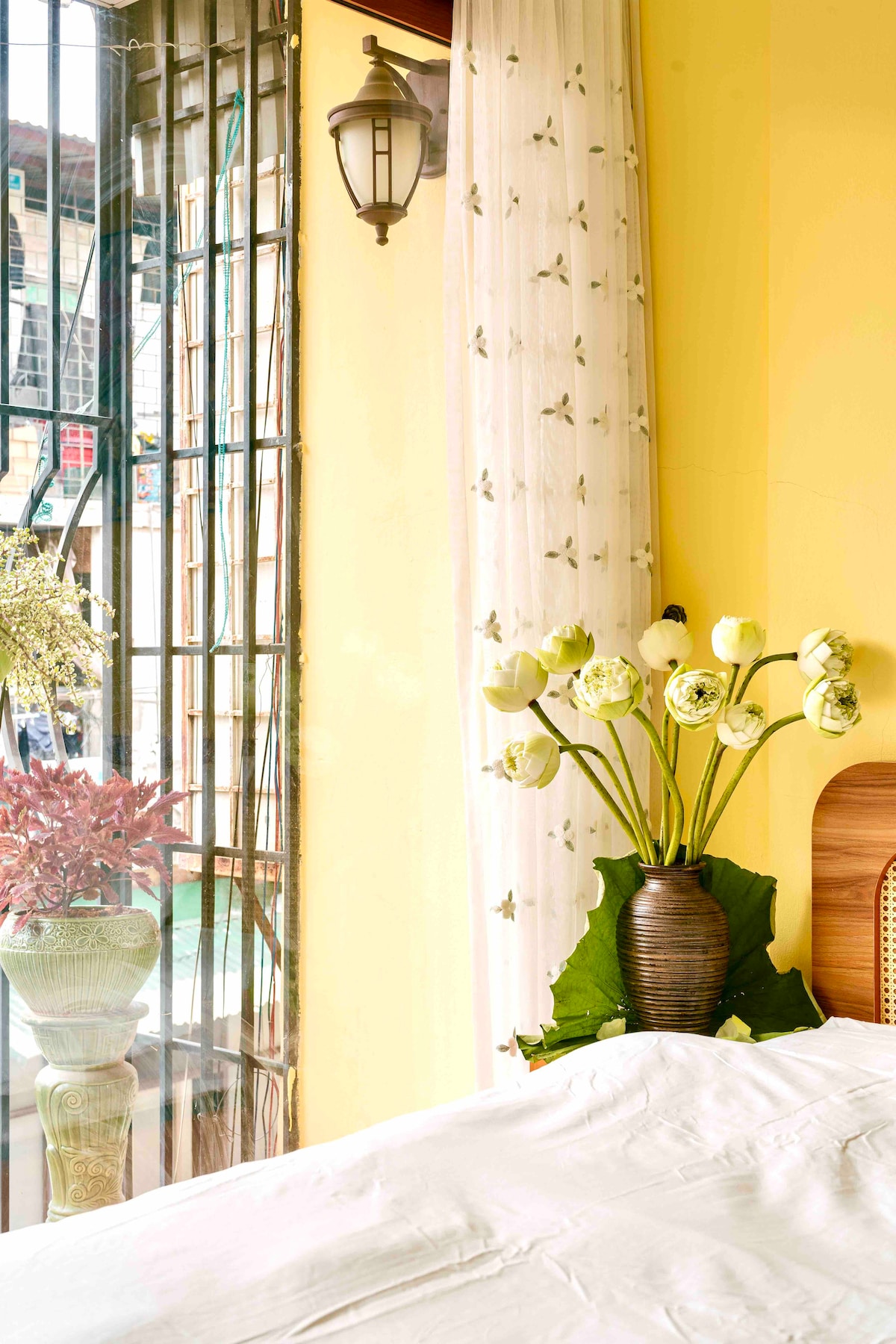 A bright yellow wall adds warmth to the space, where a cozy bed is positioned near a window. A vase of lotus flowers sits on a bedside table, complemented by lush greenery visible through the window, inviting natural light into the room.