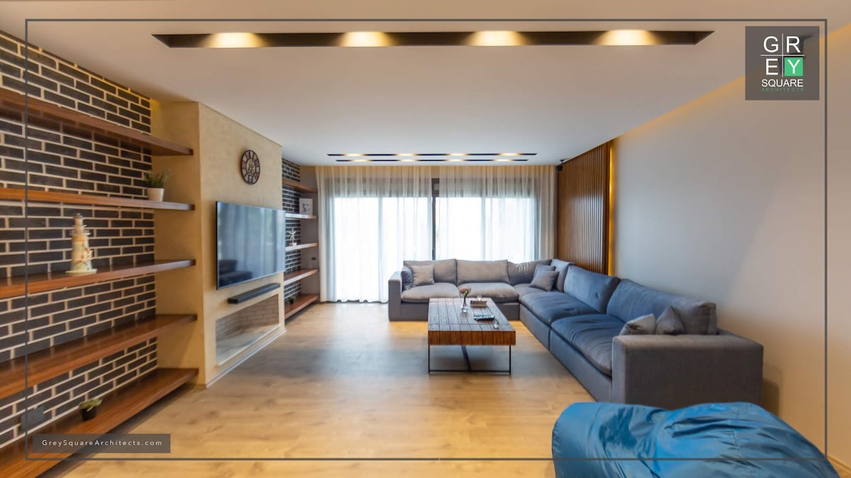 A spacious living area features a large sectional sofa arranged around a wood coffee table. A flat-screen TV is mounted on a textured wall, while open shelving displays decorative items. Natural light enters through expansive windows, draped with sheer curtains.