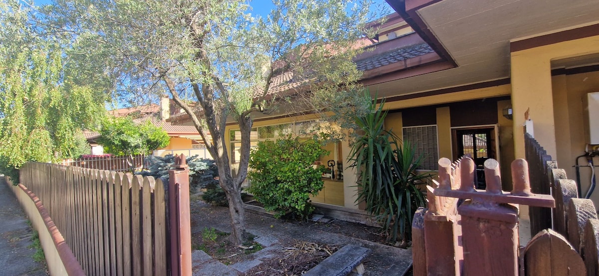 A serene exterior view of the villa is displayed, featuring a well-tended garden with green foliage and a mature tree. The façade is accented by warm tones and a tiled roof, and a wooden fence lines the pathway leading to the entrance.