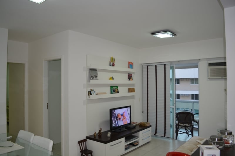 A bright living area is displayed, featuring a flat-screen TV positioned on a low, modern console. White walls are complemented by shelving with decorative items. Light is provided by ceiling fixtures, and large glass doors lead to a balcony with seated outdoor furniture.