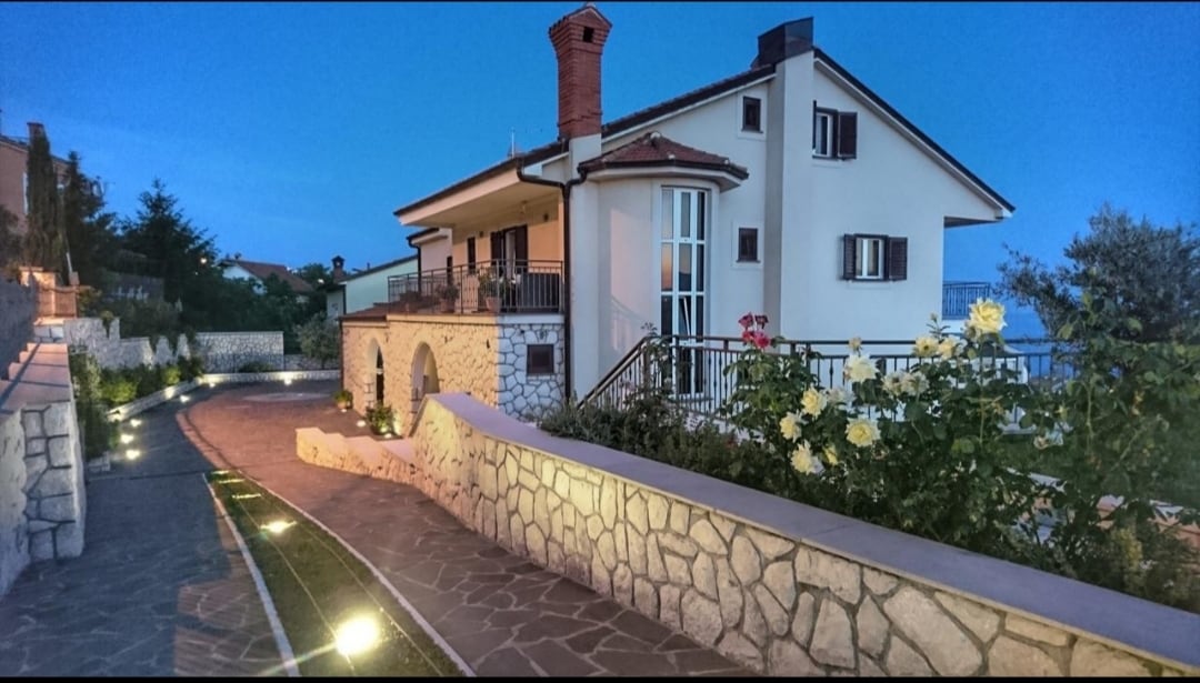 The exterior of the house is presented at dusk, showing a two-story structure with a prominent chimney and several windows. A stone pathway bordered by lush gardens leads to the entrance, illuminated by soft outdoor lighting, creating a welcoming approach.