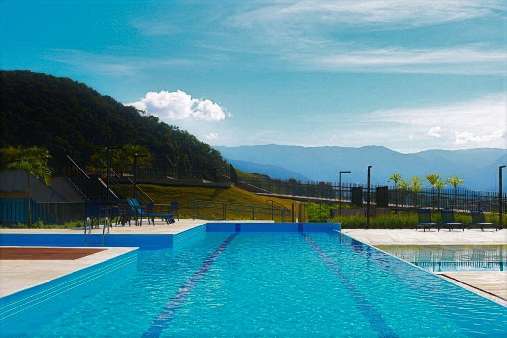 A view of a sparkling swimming pool reflects the sky and surrounding mountains. Lounge chairs line the poolside, providing a space for relaxation. Lush greenery is visible in the background, contributing to a serene outdoor environment.