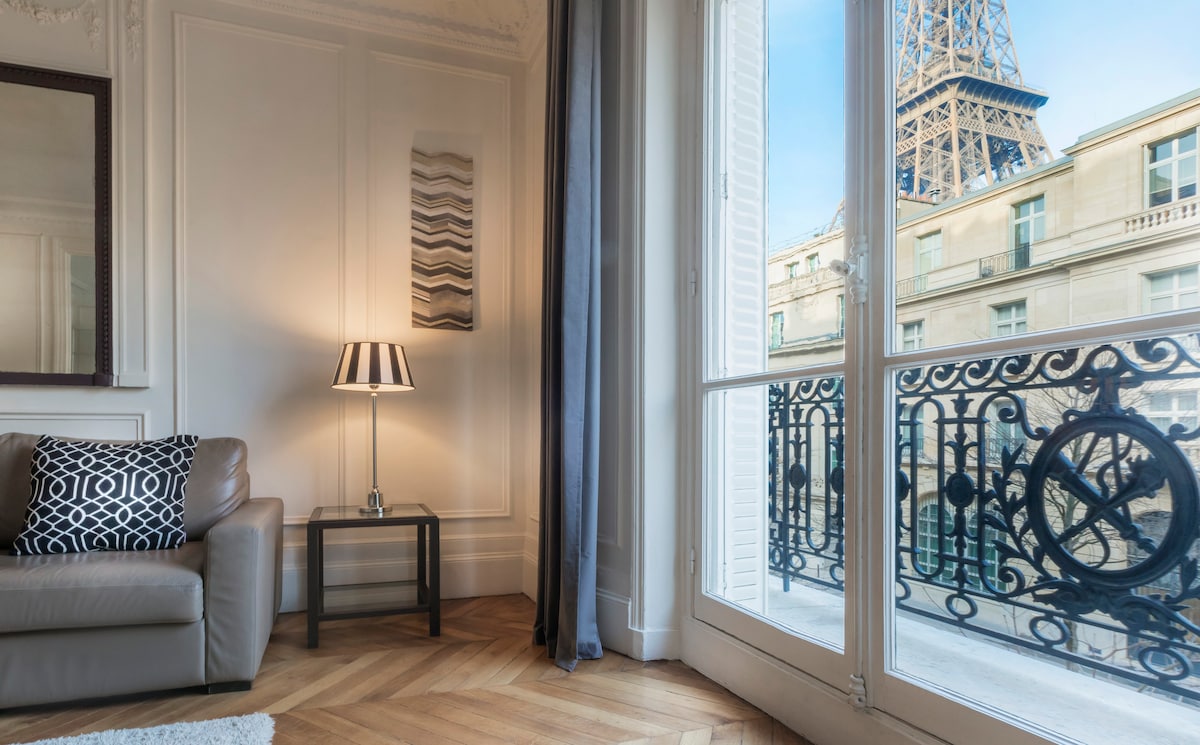 A corner of the living area features a comfortable sofa beside a side table with a lamp. Large windows offer a view of the Eiffel Tower, framed by elegant balcony railings. Light wood flooring enhances the sophisticated ambiance.