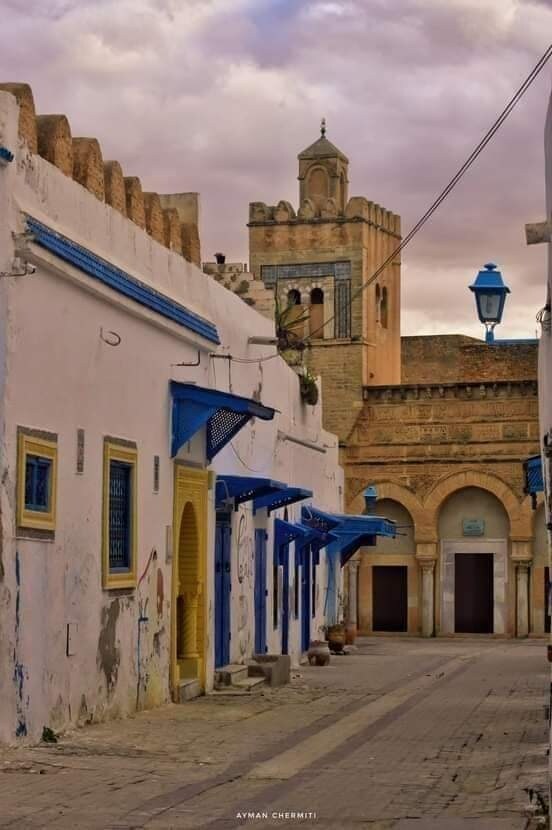 A narrow street features whitewashed buildings with blue doors and window frames, showing a blend of architectural styles. An ancient stone tower and a decorative lantern are visible in the background, while the cobblestone pavement creates a charming atmosphere.