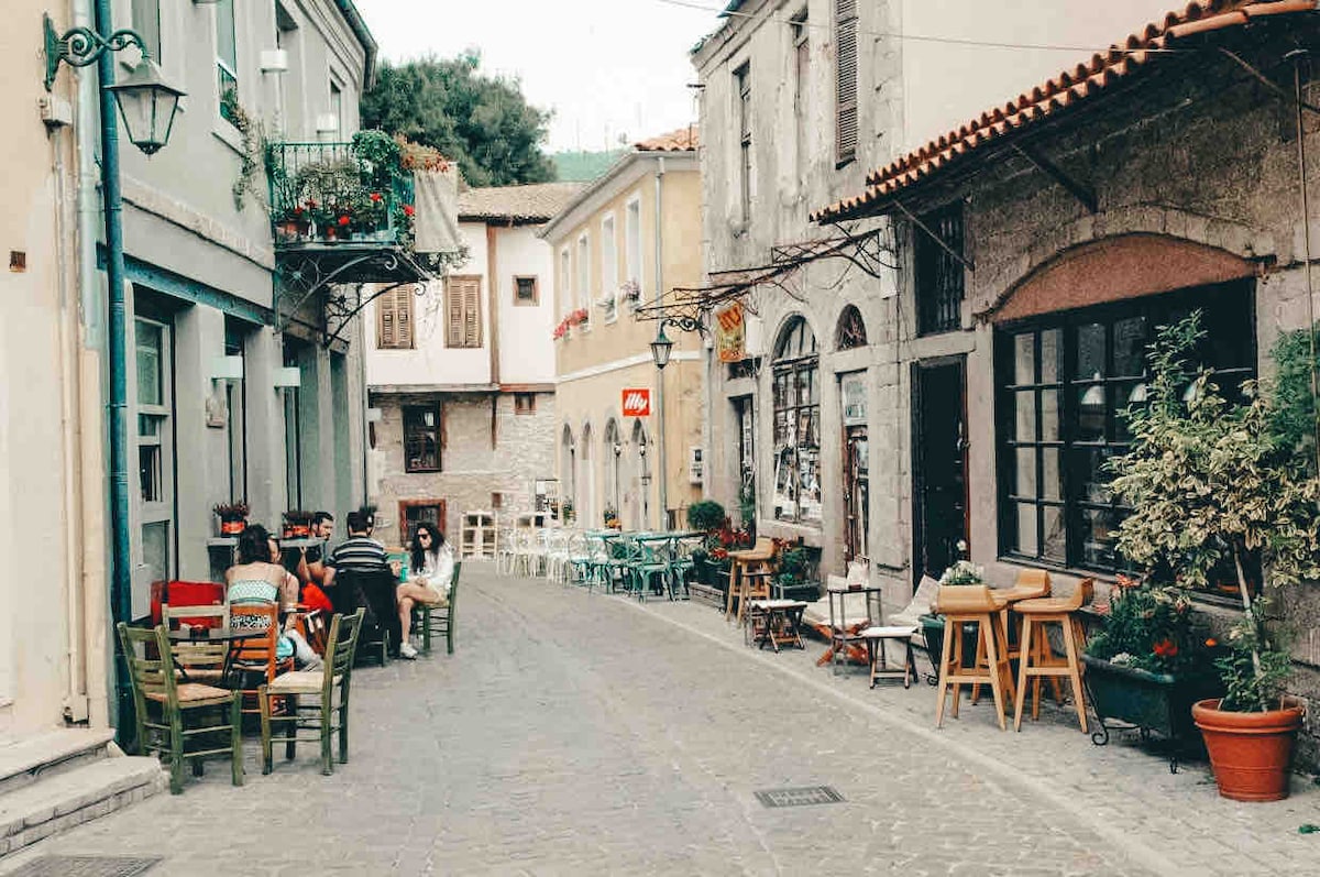 A charming cobblestone street is lined with a mix of outdoor seating and small tables, shaded by nearby buildings. Colorful chairs and potted plants add a vibrant touch. The architecture features traditional stone walls and wooden accents, creating an inviting atmosphere for casual dining.