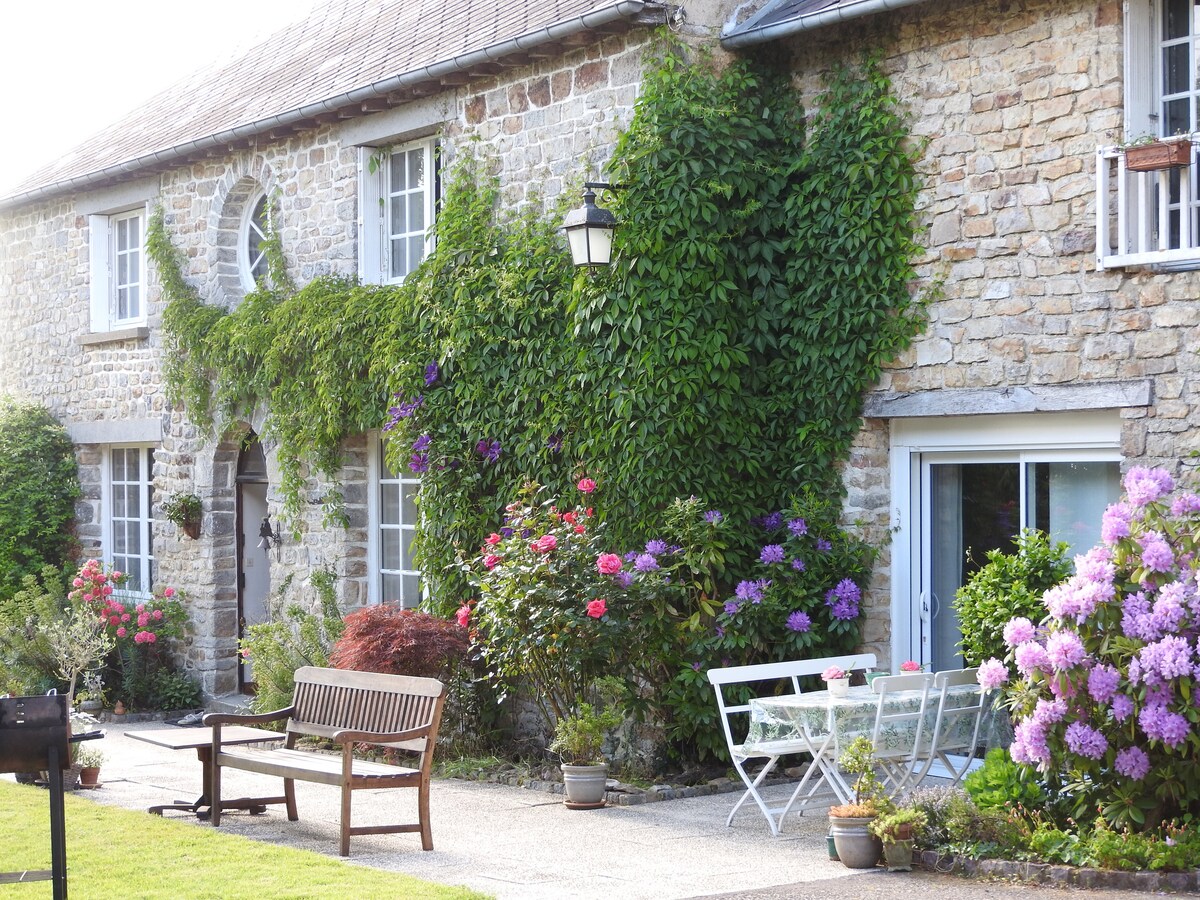 A charming stone exterior of a house is surrounded by lush green ivy and colorful flowering plants. A wooden bench and a small table with chairs are positioned in the front yard, creating an inviting outdoor space enhanced by seasonal blooms.
