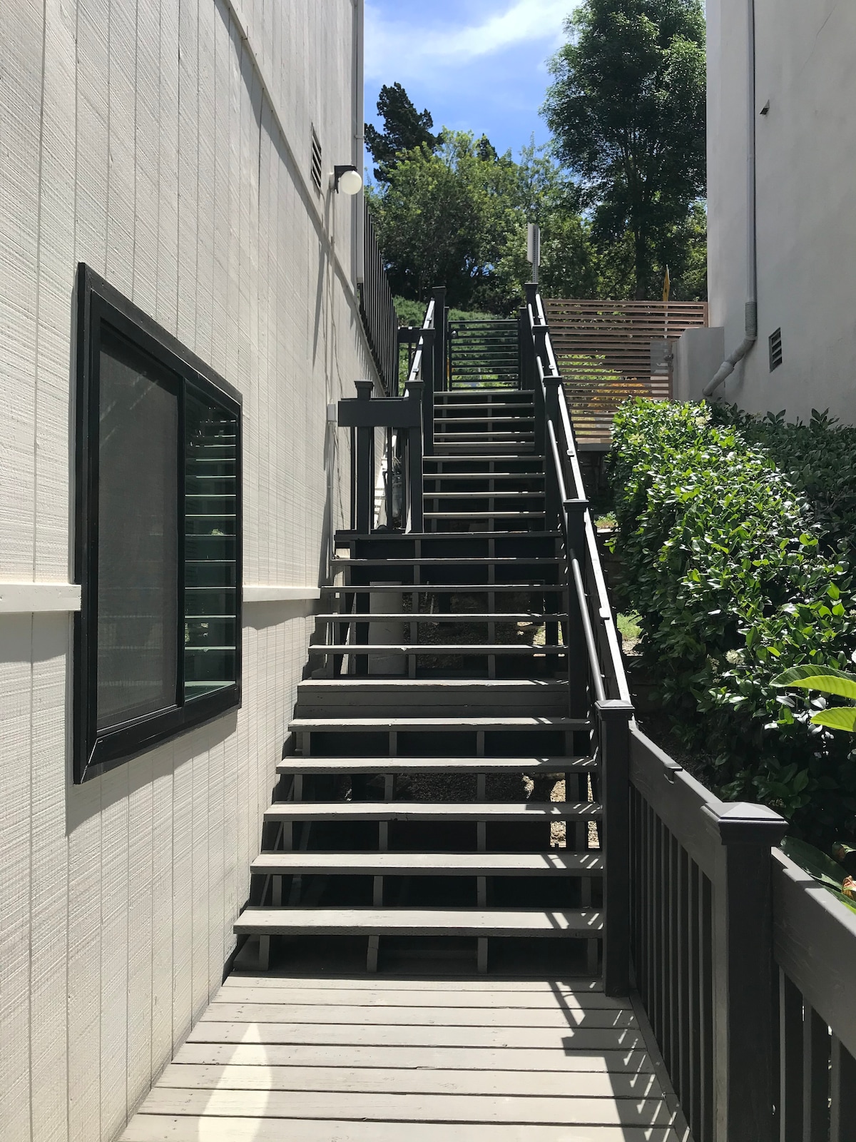A private stairwell is shown, featuring wooden steps leading upward to the entrance of the main floor. The walls are painted in a light tone, and greenery is visible on either side of the staircase, contributing to a serene outdoor environment.
