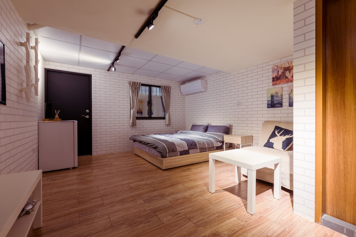 A spacious room is highlighted by a large bed with neutral bedding, complemented by a seating area featuring a light-colored sofa and a simple coffee table. Soft natural light enters through a large window framed by curtains, and a mini fridge is placed near the door.