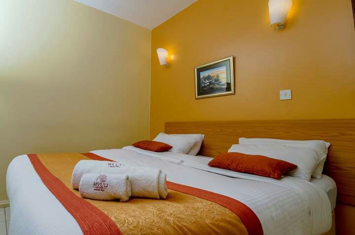 A well-lit room features two spacious beds with decorative pillows. A towel is neatly arranged on the bedding. An artwork hangs on the wall, and soft wall sconces provide additional illumination, enhancing the warm color palette of the room.