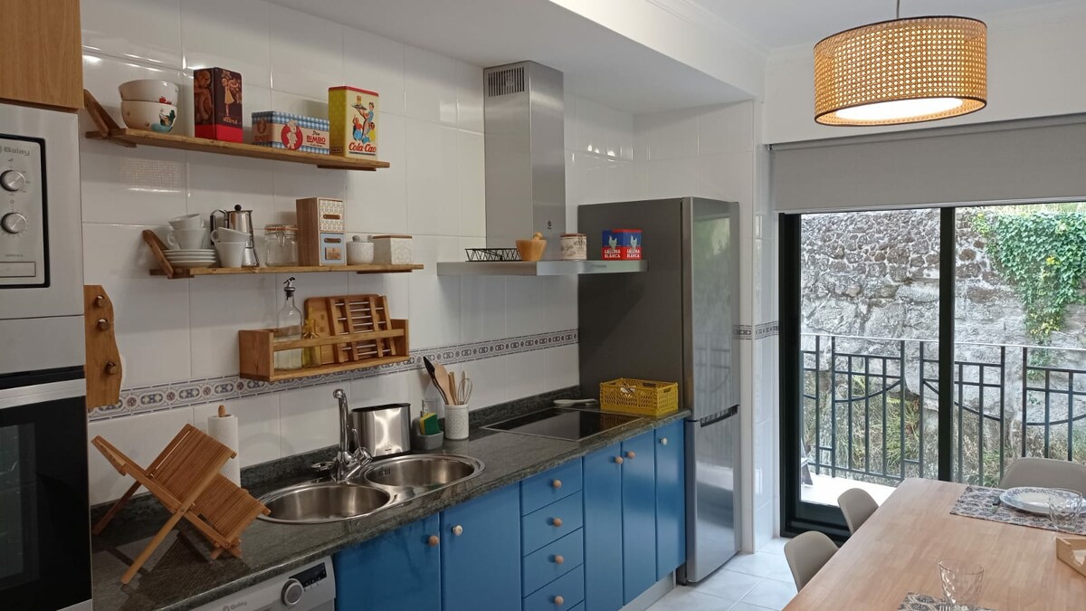 A functional kitchen with bright blue cabinetry and granite countertops is visible. Stainless steel appliances, including a refrigerator and a range hood, are present. Open shelving displays various dishware. A wooden dining table with chairs is positioned in front of large glass doors leading to a balcony.