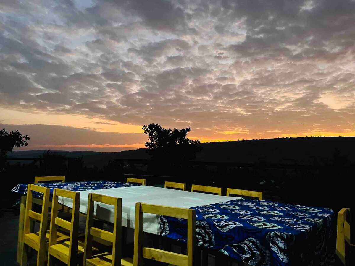 A wooden dining table is set outdoors, surrounded by matching chairs, with a traditional blue and white patterned tablecloth. The sky is filled with clouds, displaying soft hues of orange and purple as the sun sets over a distant horizon, providing a serene backdrop.