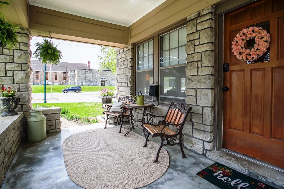 The front porch features a round area rug on a concrete floor, complemented by two wrought iron chairs and a small table. A welcoming door adorned with a floral wreath provides access, while hanging plants and potted flowers add natural touches beside the entrance.