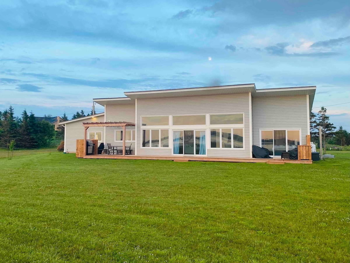 A modern beach home is depicted with a spacious deck and expansive glass doors, allowing ample natural light. The structure is set on a green lawn, surrounded by trees, with outdoor seating visible on the deck. The serene exterior reflects a contemporary architectural style.