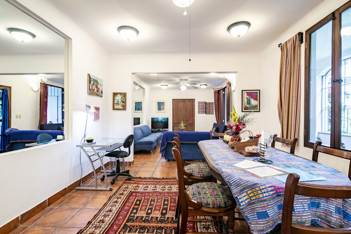 A dining area features a large table surrounded by wooden chairs, complemented by a colorful patterned tablecloth. A workspace is positioned to one side, and soft seating in blue tones is visible in the background. Natural light streams through large windows, enhancing the spaciousness.