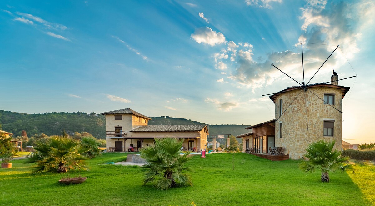 A spacious property with a charming stone windmill stands amidst a verdant landscape. Lush green grass surrounds the buildings, and the scenic hills provide a picturesque backdrop. The sky presents a soft gradient of colors with wispy clouds, enhancing the peaceful setting.