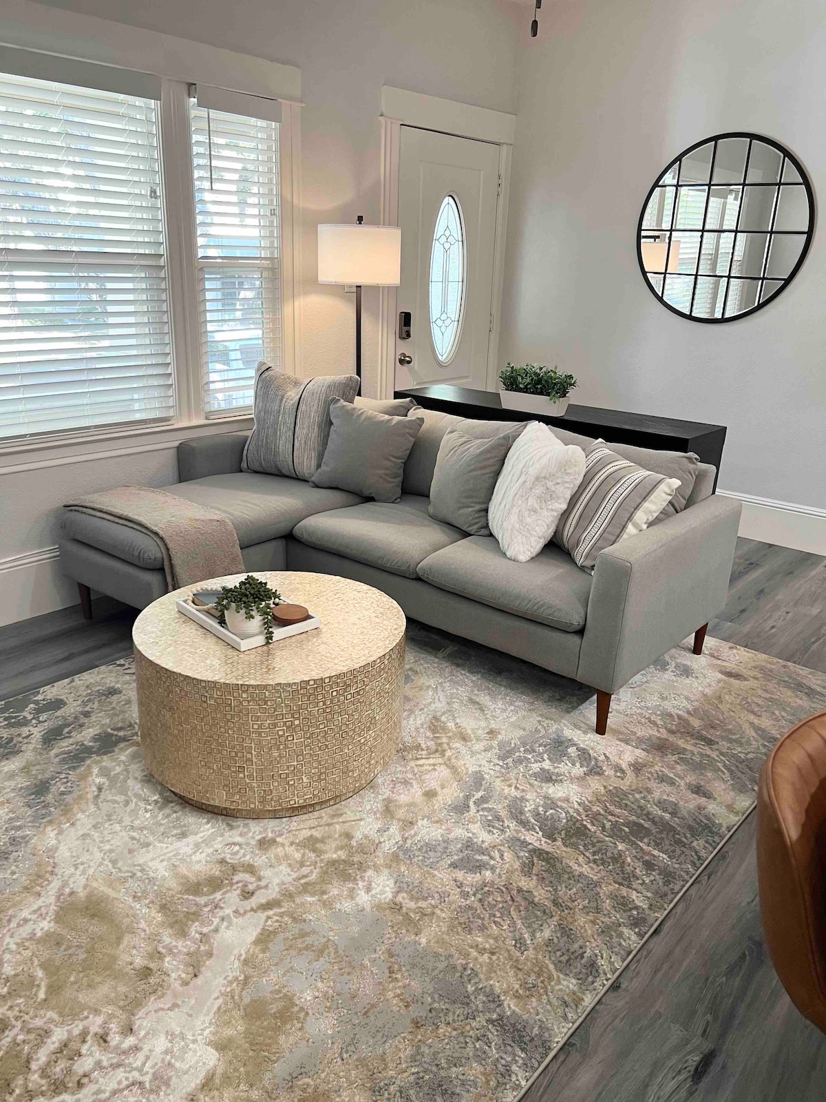A comfortable sectional sofa is arranged against a wall, accented by light-colored pillows. A round, patterned coffee table sits in front, complemented by a small decorative plant. Natural light flows in through the window, highlighting the room's neutral tones.